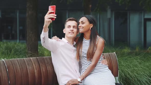 Cheerful Young Couple Taking Selfie Sitting on Bench Outdoors