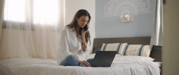 A woman talking to someone on the phone while working with a laptop from home. 