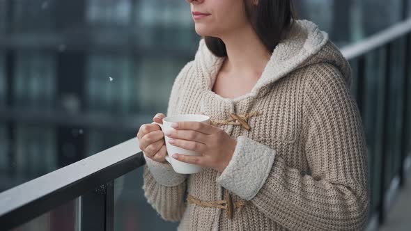 Caucasian Woman Stays on Balcony During Snowfall with Cup of Hot Coffee or Tea