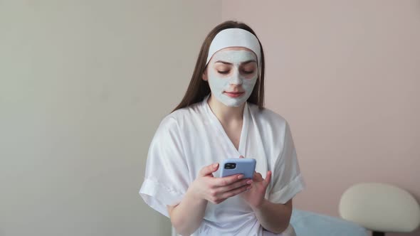 Young Woman in Beauty Cosmetology Salon with Clay Mask on Face Uses Phone