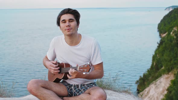 Young Smiling Man Playing Ukulele and Singing  Sitting on the Mountain and Looking in the Camera