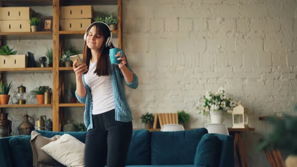 Cheerful Girl Is Dancing at Home Listening To Music Through Headphones and Holding Smartphone