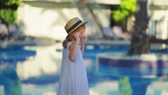 Cute Little Girl in a White Dress and Straw Hat By the Pool Near the Villa