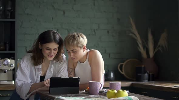 Young Hipster Attractive Young Woman Friends Watch Video Movie Surf Internet in Kitchen Using Tablet