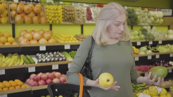 Portrait of Senior Confident Caucasian Woman Choosing Between Pomelo and Pear in Grocery and