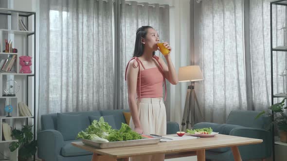 Smiling Asian Woman Enjoys Drinking Orange Juice While Preparing Healthy Food At Home