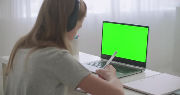 Teen Girl Is Learning Online Sitting at Table in Front of Laptop with Chroma Key on Display, Writing