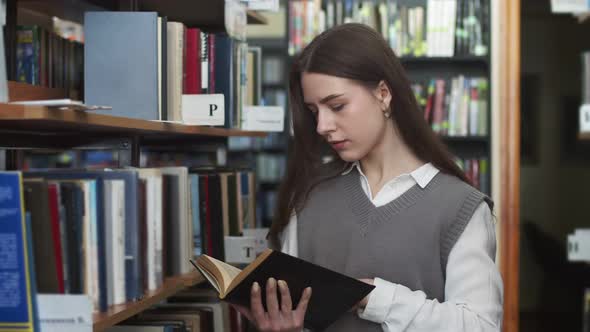 Beautiful student in corridor of library reads book standing next to the shelf. Young woman in shirt