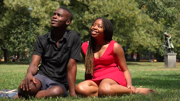A Black Man and a Black Woman Sit on Grass in a Park, Look Around and Talk