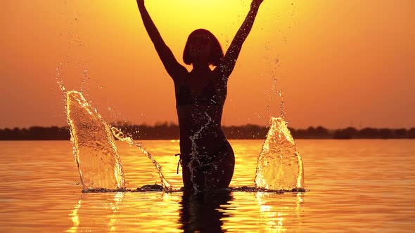 Silhouette of Woman at Sunset Raises Hands Up and Creating Splashes of Water