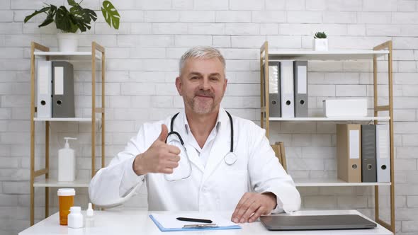 Male Doctor Gesturing Thumbs Up Sitting In Office