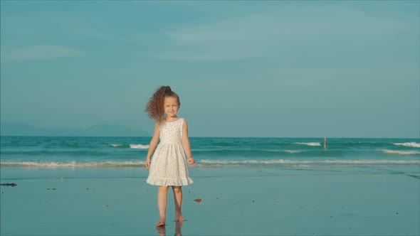 Curly Little Girl in White Dress Walking on the Beach at Sunset. Slow Motion. Happy Childhood