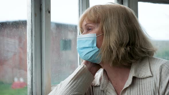 Elderly Woman In Medical Mask Looks Hopelessly Out Of Window At Self Isolation