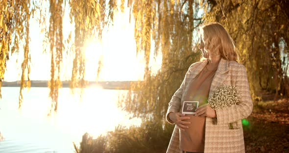 Pregnant Blonde Woman with a Bouquet on the Shore of the Pond