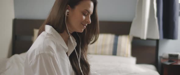 Close up of a young woman talking to someone through earphones while sitting on bed. 