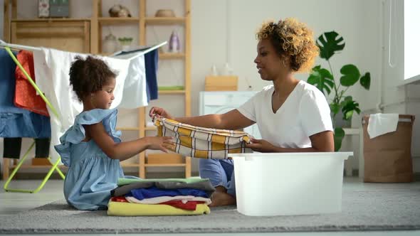 Young Mother and Little Daughter Lay Out Clothes While Sitting on Floor in Laundry at Home Spbd.