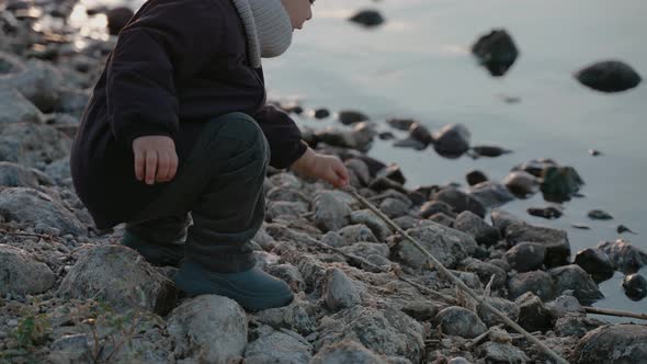 Baby Boy Picks Up a Twig to Play with on the Stone Shore on an Autumn Evening