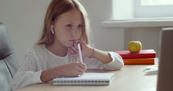Serious Focused Schoolgirl Sits at Table at Home Wears Headphones, Waving Hand Greets Online Teacher