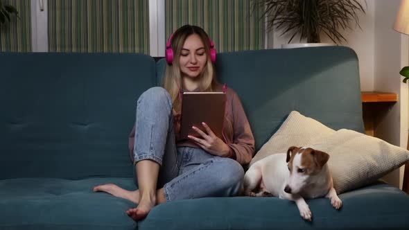 A Girl on the Couch Lies with a Dog in Her Laptop and Listens to Music Through Headphones