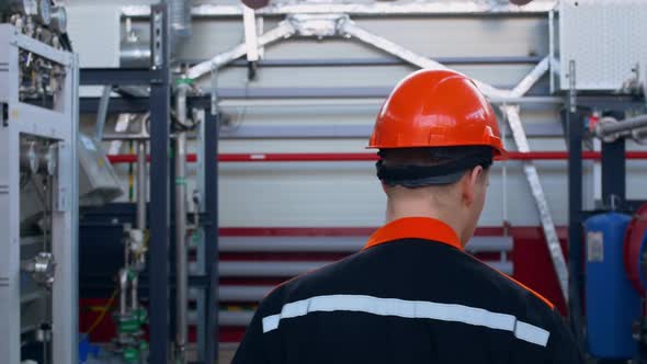 A View From the Back of a Worker in an Orange Helmet and Overalls Walks Around His Equipment at a