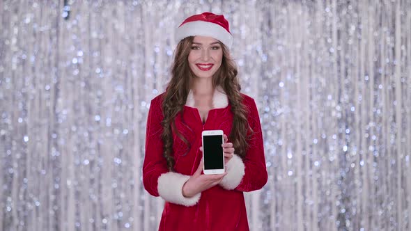 Santa Girl Holds the Phone in Her Hands and Looks Into the Distance. Bokeh Background