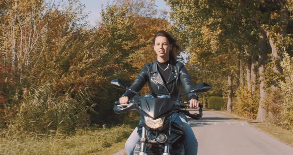 Pretty smiling European young woman driving a motorbike wearing leather jacket in forest with vibran