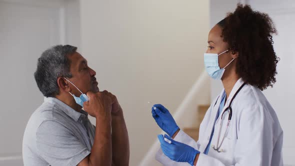 Mixed race female doctor wearing mask doing swab test on senior man at home