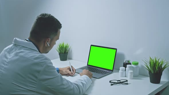 Young man doctor physician wearing white coat using laptop computer writing notes at workplace.