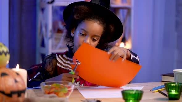 Little African American Girl in Festive Costume and Witch Hat Cuts Pumpkin Out of Orange Paper