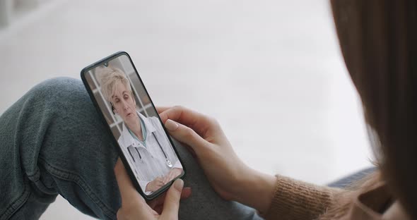 Female Medical Assistant Wears White Coat, Headset Video Calling Distant Patient on Smartphone