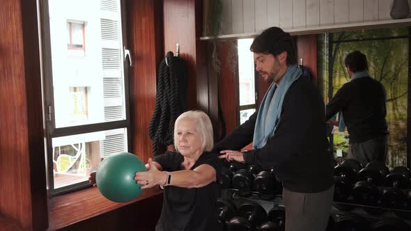 Elderly sportswoman training arms with ball near personal trainer in the gym