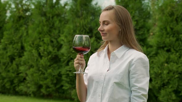 Young Woman in a White Shirt Sniffs and Tastes Red Wine on a Green Background