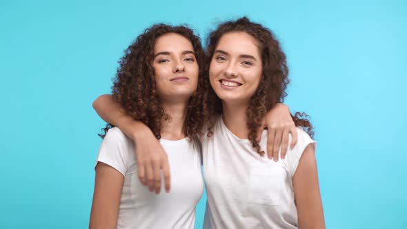 Two Charming Caucasian Curly Female Twins in White Tshirts Embracing Each Other on Shoulders and