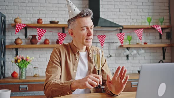 Happy Middle Aged Man in Party Cap with Champagne Glass Toasting to Laptop Camera and Wishing Good