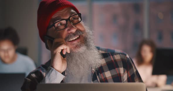 Senior Grayhaired Man in Glasses and Red Hat Talking on Mobile Phone in Office