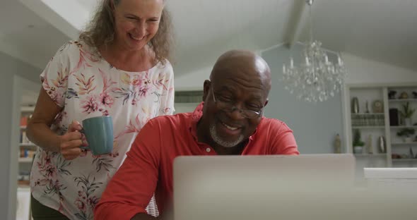 Happy senior diverse couple wearing shirts and using laptop in living room