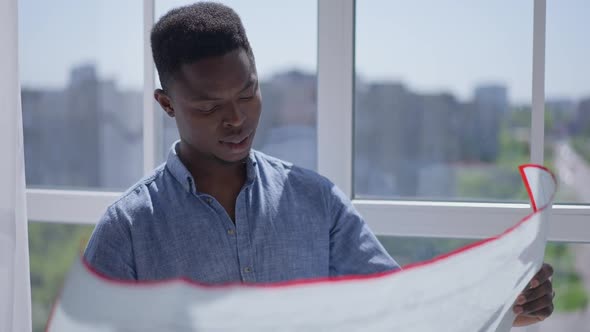 Young Creative Smart Landscaper Architect Examining Blueprint Standing at Window in Home Office