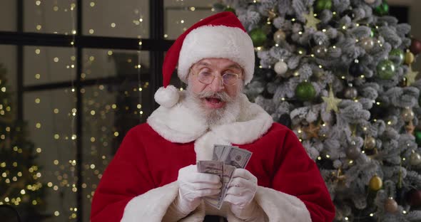 Happy Elderly Man Dressed Like Santa Claus Quickly Counts Money at Home Against the Background of a