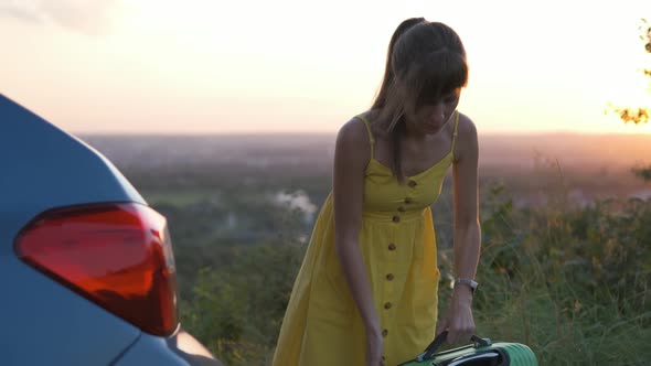 Young Woman in Yellow Dress Putting Green Suitcase in Car Trunk