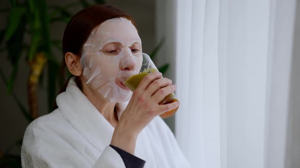 Mature Woman in Face Mask Drinking Vegetarian Smoothie Looking Out the Window Indoors