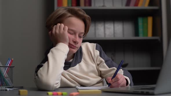 Closeup Portrait of Asleep Pupil Boy Writing in Notebook Holding Head with Hand Sitting on Desk