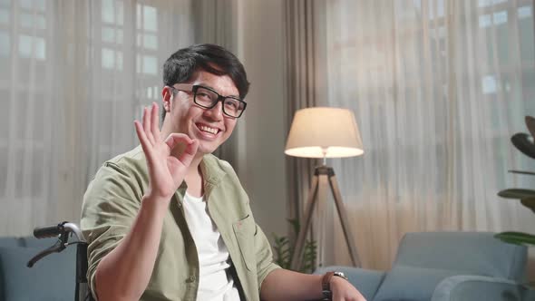 Asian Man Sitting In A Wheelchair Smiling And Showing Ok Sign In Living Room