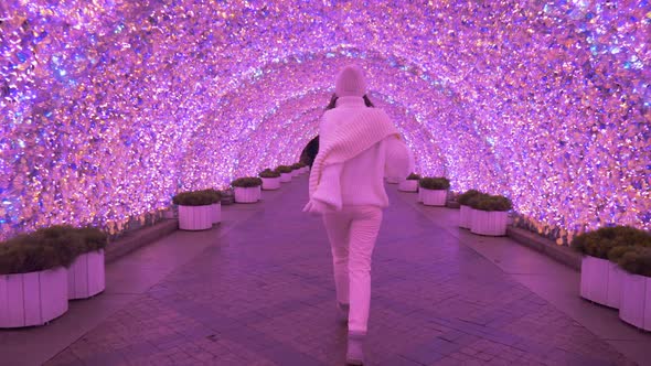 Woman in a White Sweater and Hat Walks Through a Pink Glowing Tunnel at Night