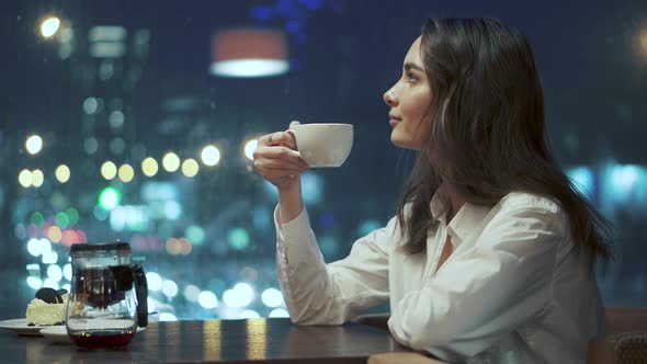 Girl Drinks Tea and Admires the View of the Night City Sitting in a Cozy Cafe Late in the Evening