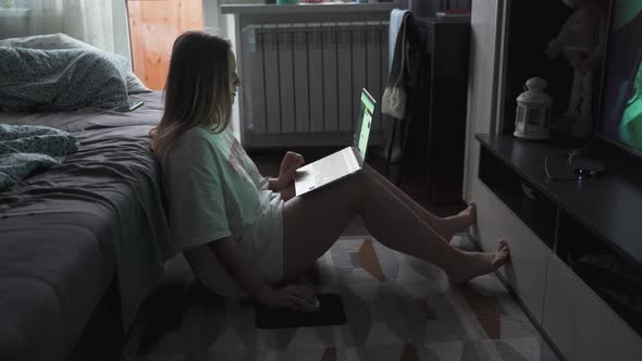 Young Woman is Sitting on the Floor on Carpet in Front of the TV and Working on Laptop in the Early