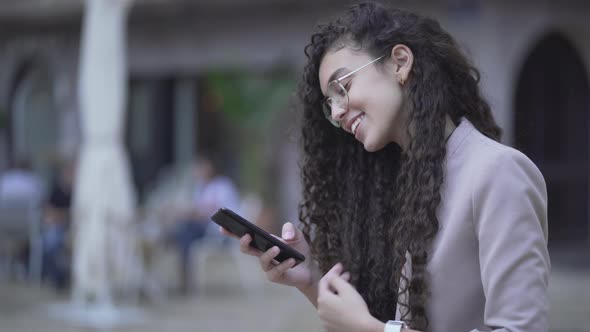 Moroccan Girl With Black Curly Hair Smiling While Texting On Smartphone