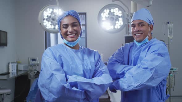 Portrait of diverse male and female surgeon wearing lowered face masks smiling in operating theatre