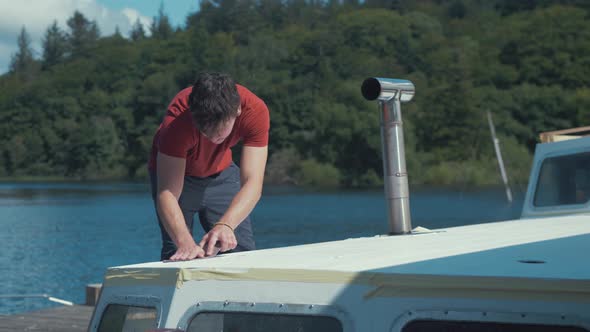 Young man sanding roof planks on wooden boat. MEDIUM WIDE SHOT