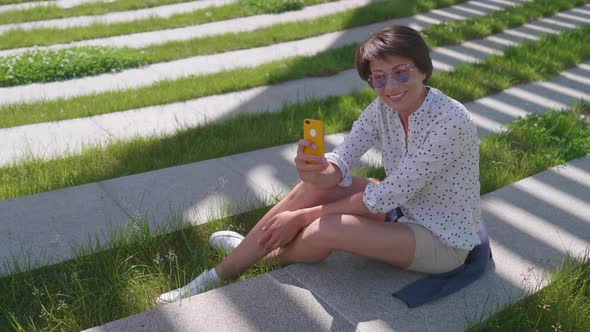 Woman with Colorful Sunglasses Sits on Lawn in Urban Park and Makes Selfie on Smartphone