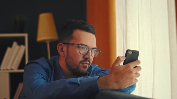 Young Arab Man at Home on the Couch Using the Phone and Looking Out the Window